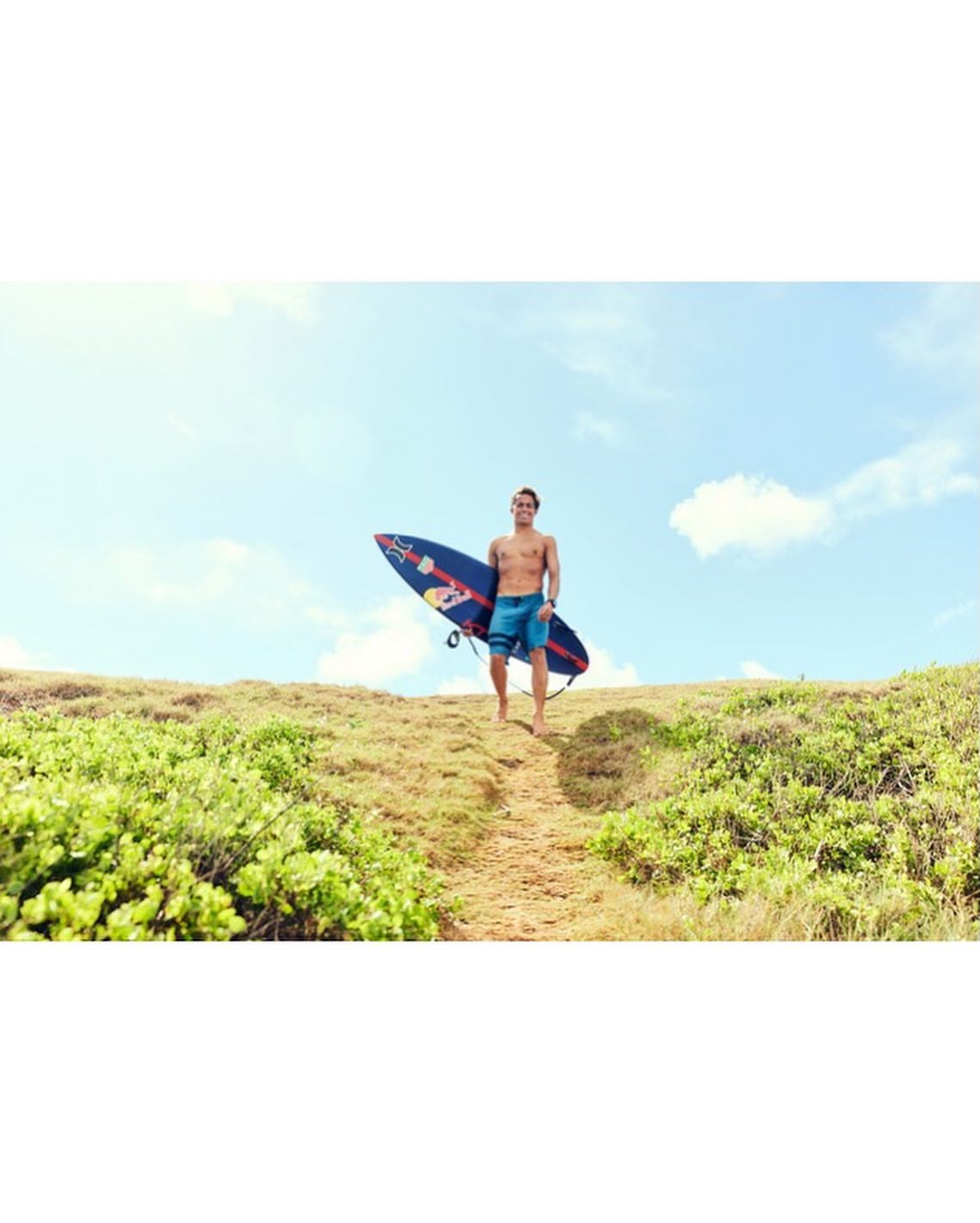 Shirtless male surfer carries blue surfboard up grassy hill on sunny day, blue sky with clouds, outdoor setting, medium shot.