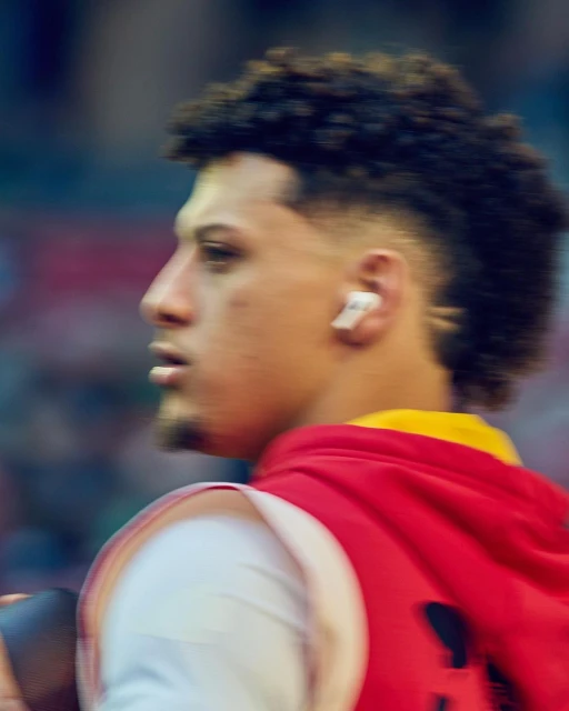Profile shot of a male athlete with a short mohawk hairstyle, wearing a red and yellow jacket, and sporting wireless earbuds.