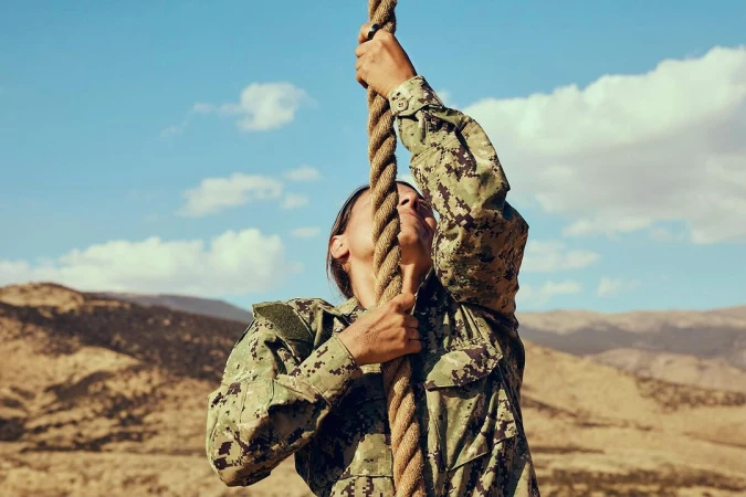 A woman in a camouflage uniform climbs a thick rope against a backdrop of a sunny sky and a tan, hilly landscape.