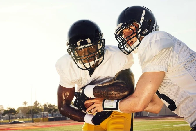 Two athletic American football players wearing helmets and pads compete on a vibrant green field, under bright sunlit conditions.