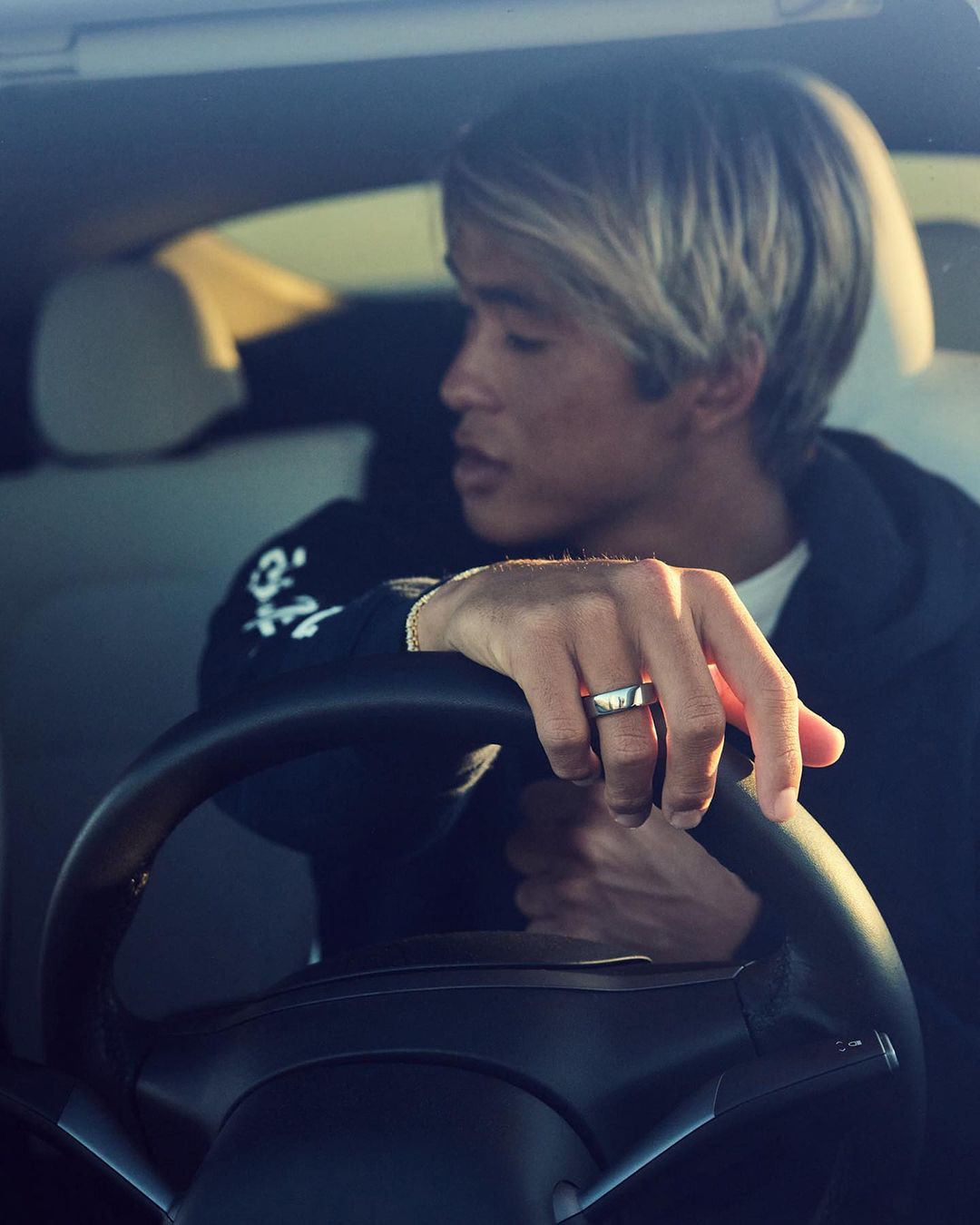 An Asian male with light blonde hair is driving a car, his hands gripping the steering wheel. He is wearing a black sweatshirt.