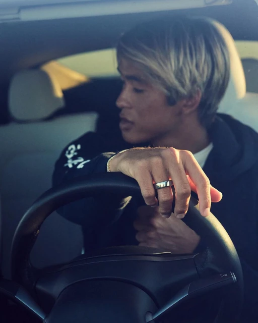 An Asian male with light blonde hair is driving a car, his hands gripping the steering wheel. He is wearing a black sweatshirt.