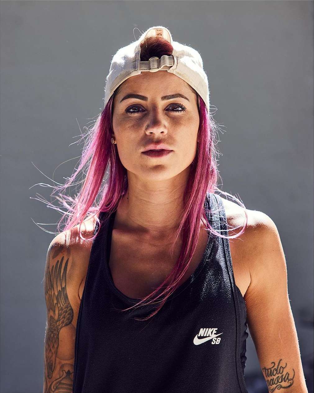 Portrait of a woman with pink hair wearing a white baseball cap and black tank top, looking directly at the camera with a neutral expression.