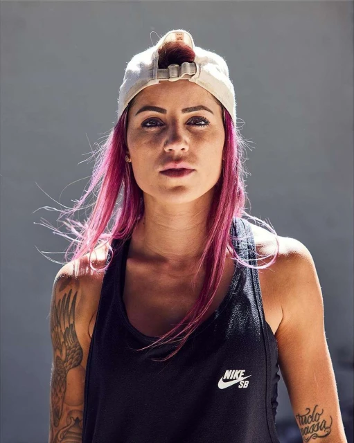 Portrait of a woman with pink hair wearing a white baseball cap and black tank top, looking directly at the camera with a neutral expression.
