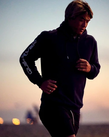 Man wearing a dark hooded sweatshirt with lettering on the sleeve running outdoors during sunset, captured by Ryan Young.