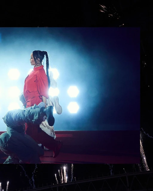 Female pop singer Rihanna is seen on stage in a red jumpsuit. Spotlight and stage lights illuminate her at a concert.