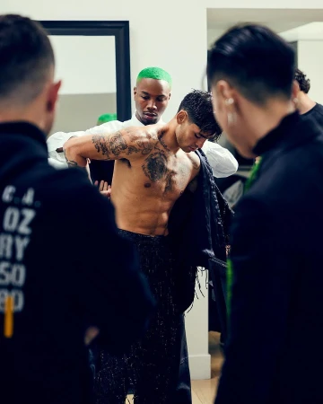 Behind-the-scenes portrait of a shirtless tattooed man being dressed by assistants in a studio setting by Ryan Young.