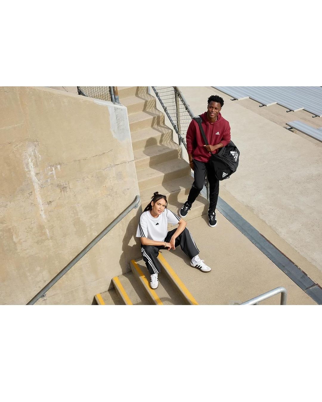 Two young adults, one seated, one standing, outdoors on concrete stairs, wearing Adidas apparel. The man carries a black Adidas sports bag.