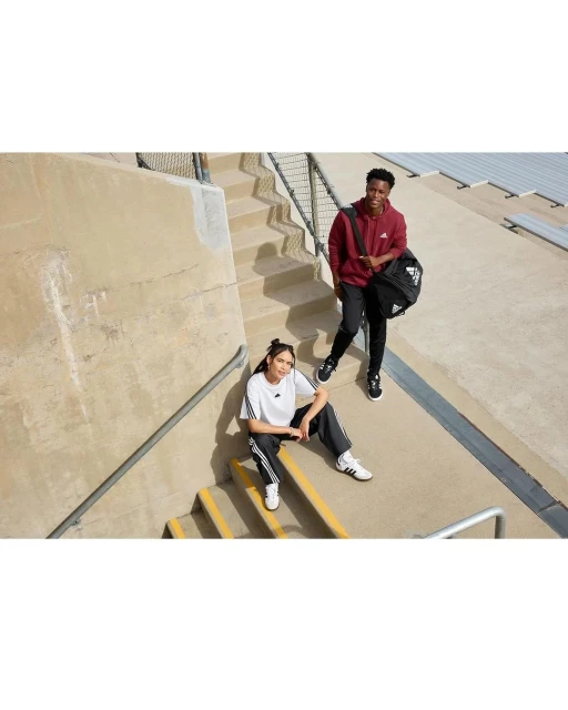 Two young adults, one seated, one standing, outdoors on concrete stairs, wearing Adidas apparel. The man carries a black Adidas sports bag.