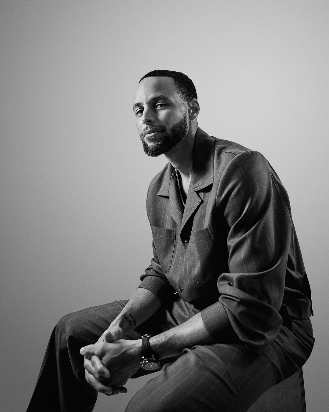 Black and white portrait of a man with a beard wearing a pinstripe button-down shirt, seated with hands clasped.