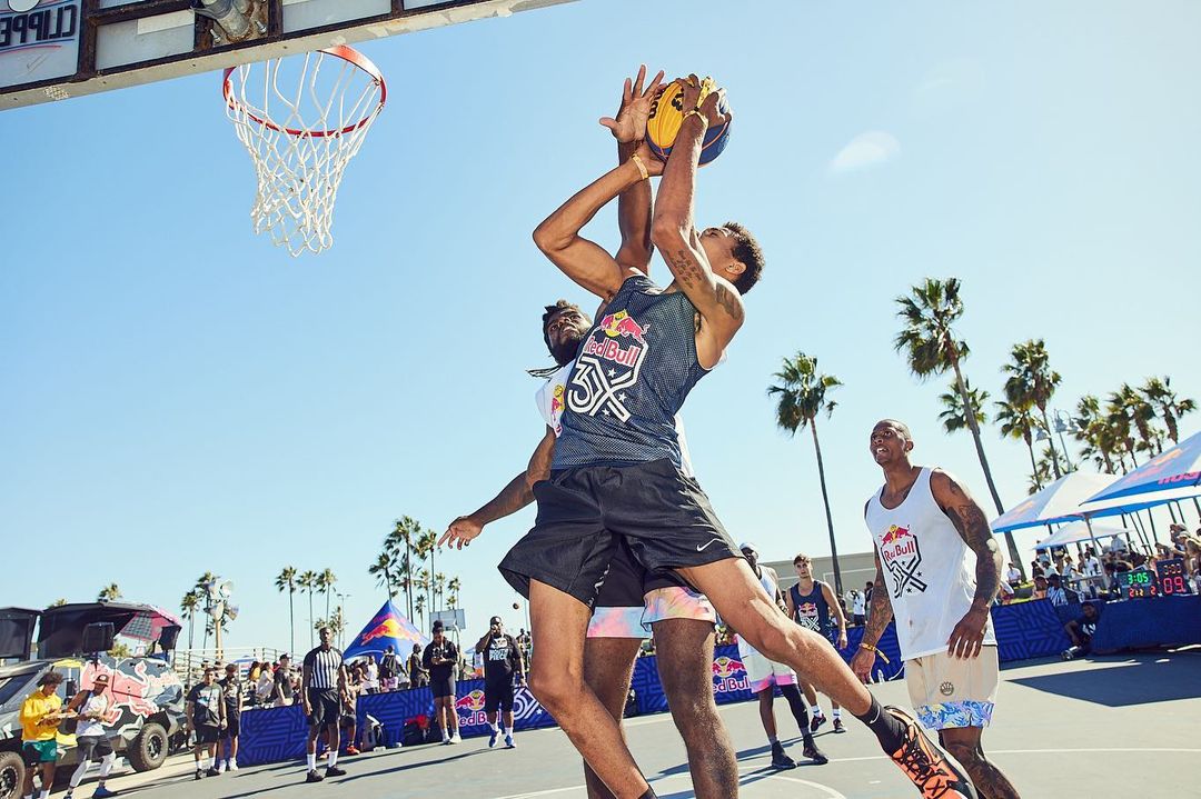 Two adult male basketball players compete for a yellow basketball during an outdoor game on a blue court in the daytime under a blue sky.