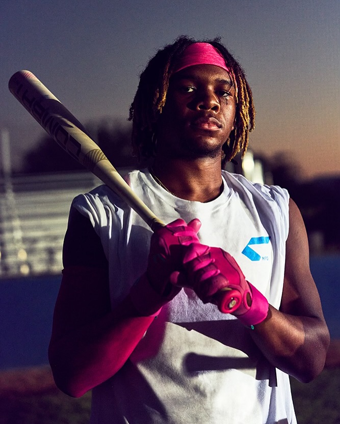 Portrait of a young Black man wearing a pink headband and gloves, holding a baseball bat in front of a baseball field.