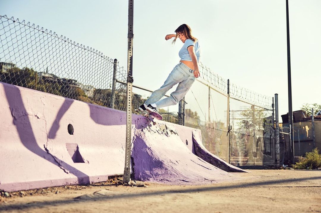 A female skateboarder is catching air over a ramp outdoors on a sunny day. The ramp is painted purple and set against a chain-link fence.
