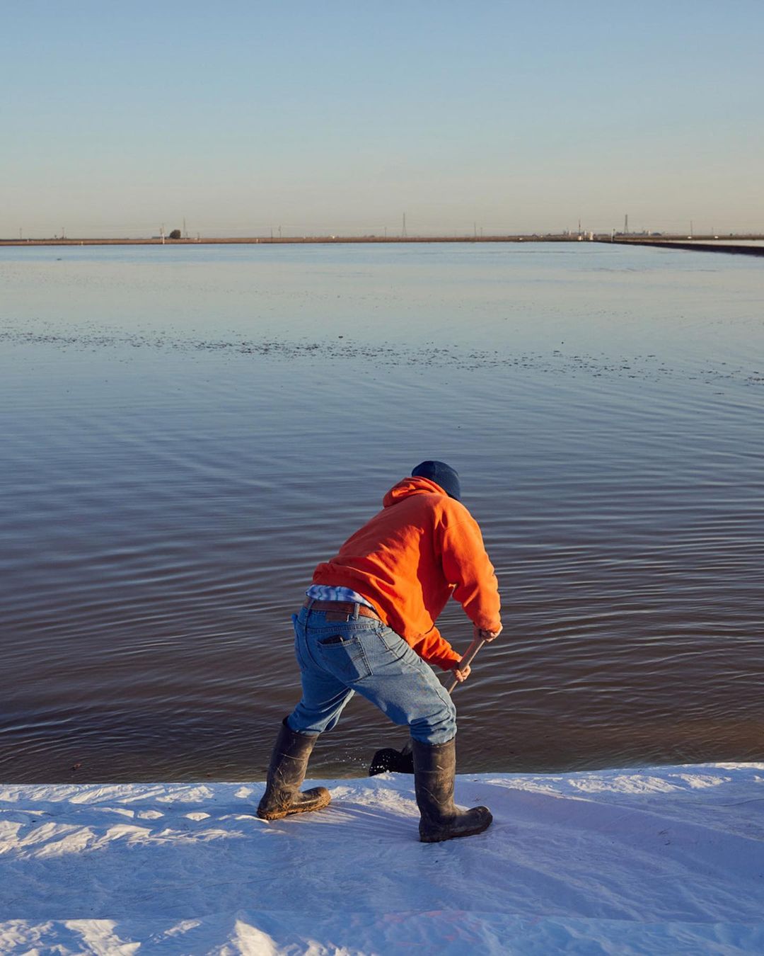 Man in an orange hoodie standing near a large body of water, wearing boots and a blue cap, working with a tool.