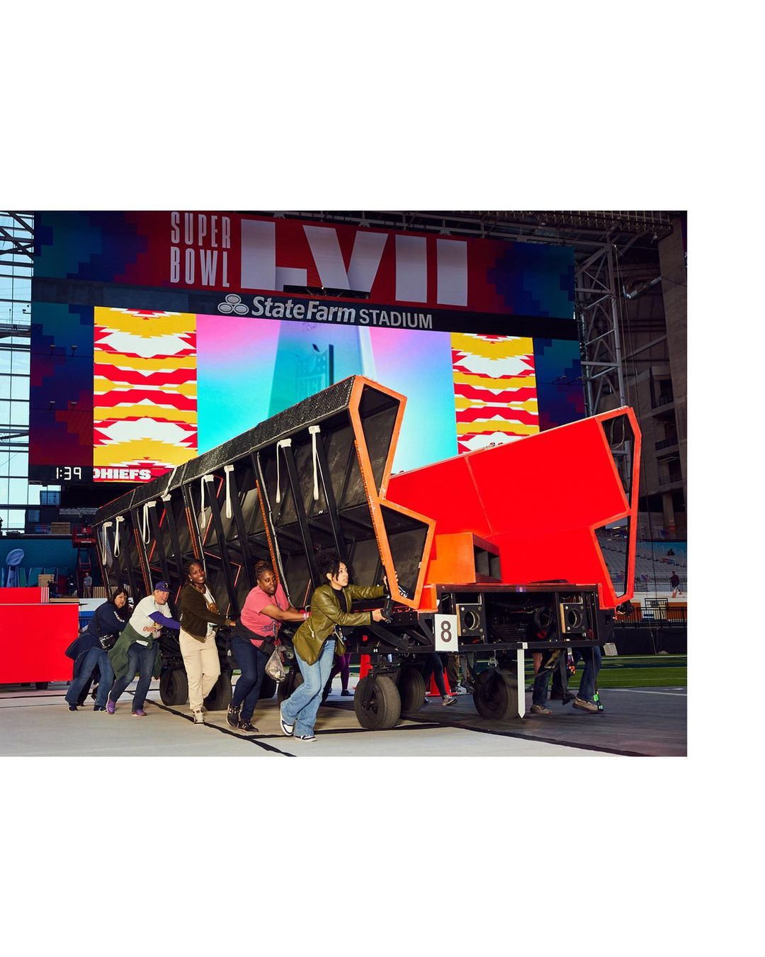 A group of people push a large black and red object on wheels in front of a Super Bowl LVII sign inside State Farm Stadium.