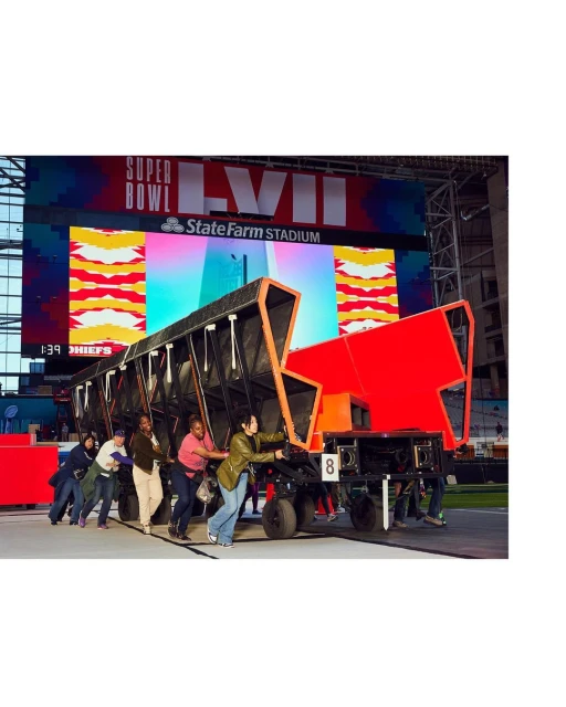 A group of people push a large black and red object on wheels in front of a Super Bowl LVII sign inside State Farm Stadium.