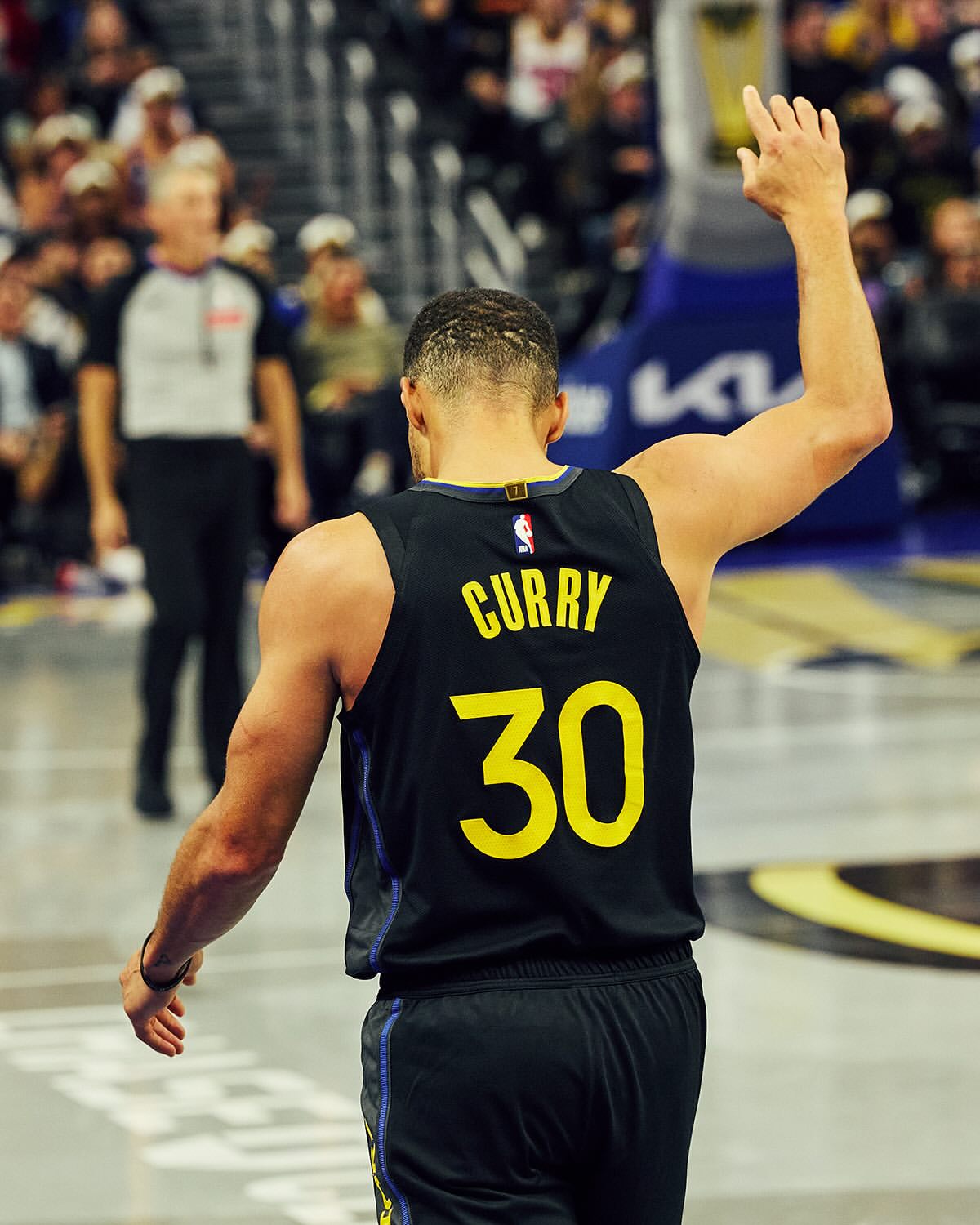 Steph Curry on the court in a Golden State Warriors jersey. He is facing away from the camera and waving, and a referee is visible in the background.