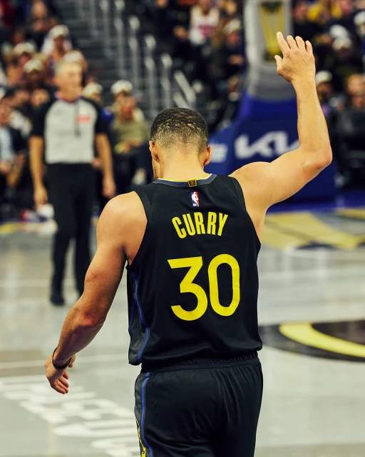 Steph Curry on the court in a Golden State Warriors jersey. He is facing away from the camera and waving, and a referee is visible in the background.