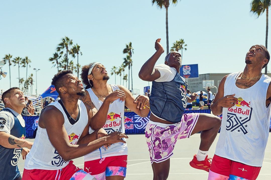 A group of male basketball players in matching jerseys look up at the basketball during an outdoor game on a sunny day.