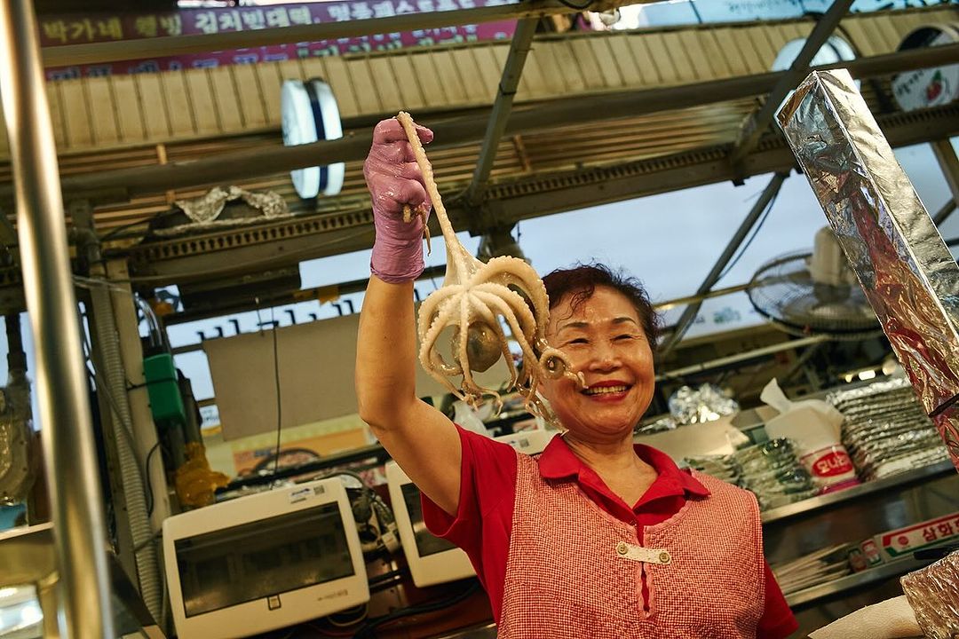 A smiling woman in a market holds up an octopus in a gloved hand, showcasing fresh seafood, with background market details.