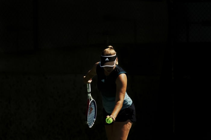 Adult female tennis player prepares to serve the ball on court in athletic wear, wearing visor with an adidas logo. The background is dark.