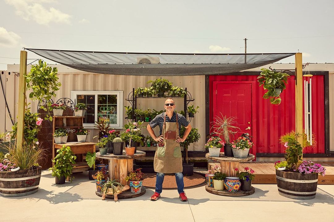 A confident man in an apron poses in front of a plant shop made from storage containers, showcasing a diverse collection of potted plants.