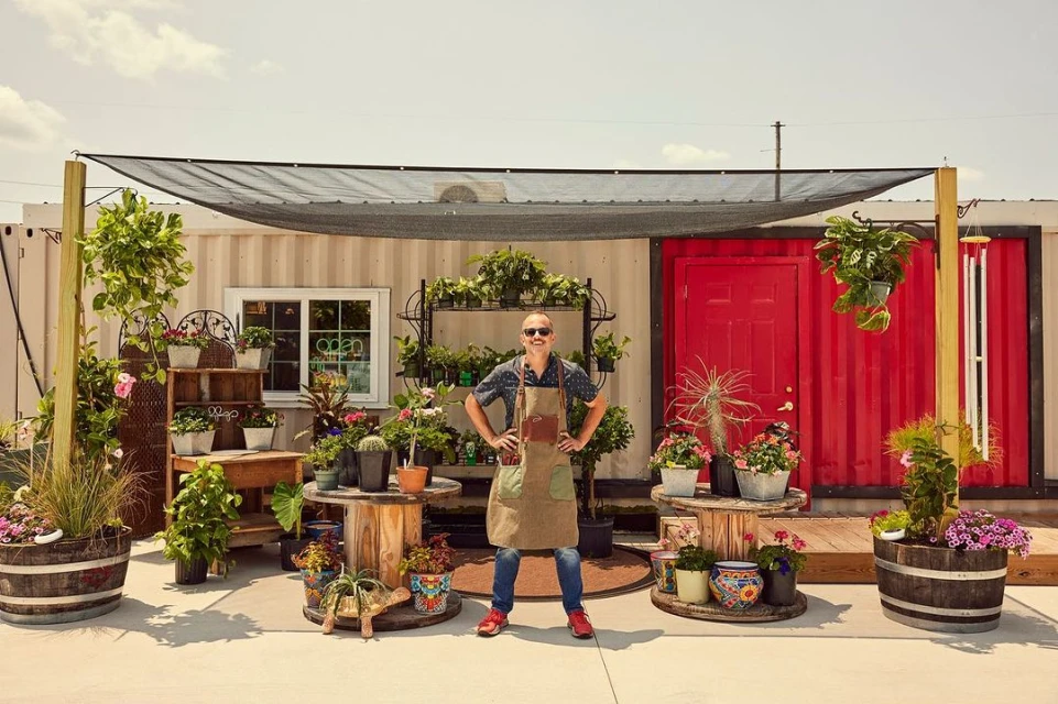 A confident man in an apron poses in front of a plant shop made from storage containers, showcasing a diverse collection of potted plants.