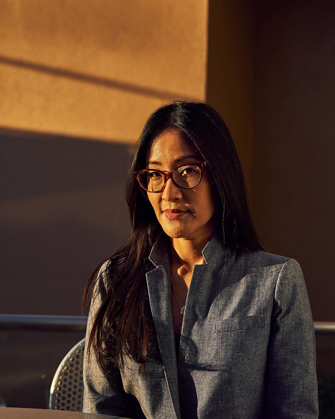 Attractive Asian woman with glasses and a blazer, sitting at an outdoor cafe or patio with an ochre-colored wall in the background.