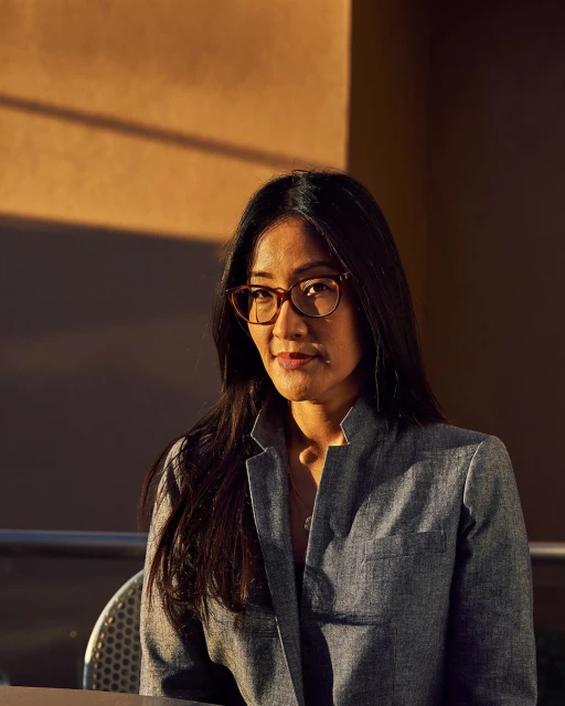 Attractive Asian woman with glasses and a blazer, sitting at an outdoor cafe or patio with an ochre-colored wall in the background.