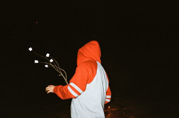 Person in hoodie holding stick with marshmallows in the dark, illuminated by a fire. Nighttime campfire snack concept.