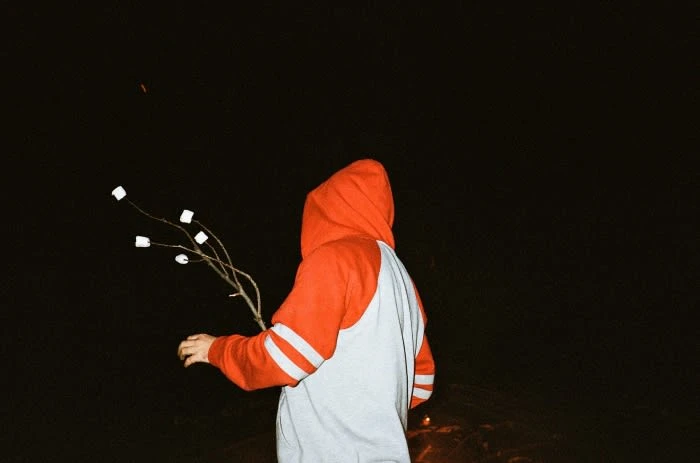 Person in hoodie holding stick with marshmallows in the dark, illuminated by a fire. Nighttime campfire snack concept.