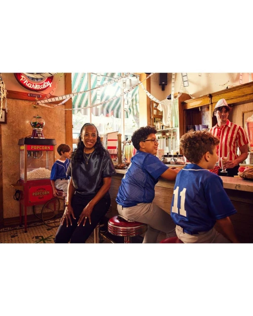 A woman and three kids at a diner counter, with a man in a server uniform. Retro diner scene with popcorn machine and vintage signs.