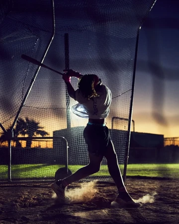 Baseball player swinging bat during sunset, wearing athletic clothing, with a netting fence and desert landscape in the background.