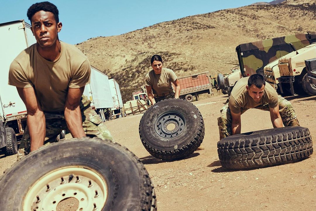Three people in military fatigues lift tires as part of outdoor fitness training in a desert environment on a sunny day.