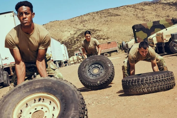 Three people in military fatigues lift tires as part of outdoor fitness training in a desert environment on a sunny day.