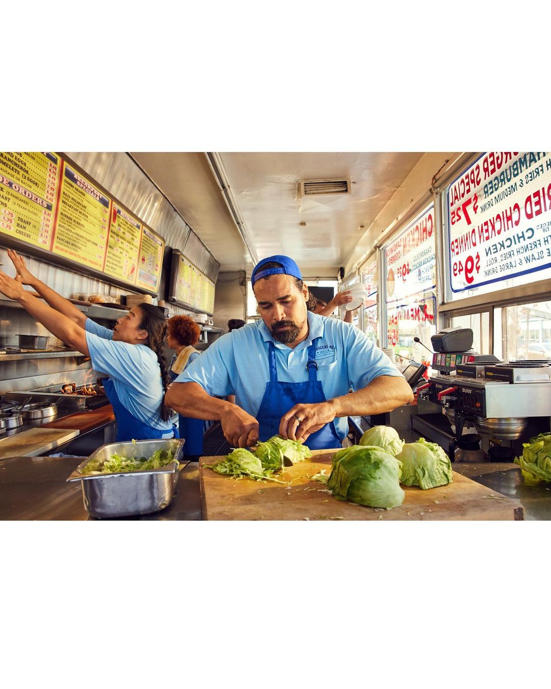 Chef at a food truck skillfully prepares lettuce for a burger, with another employee working in the background and food menu visible.