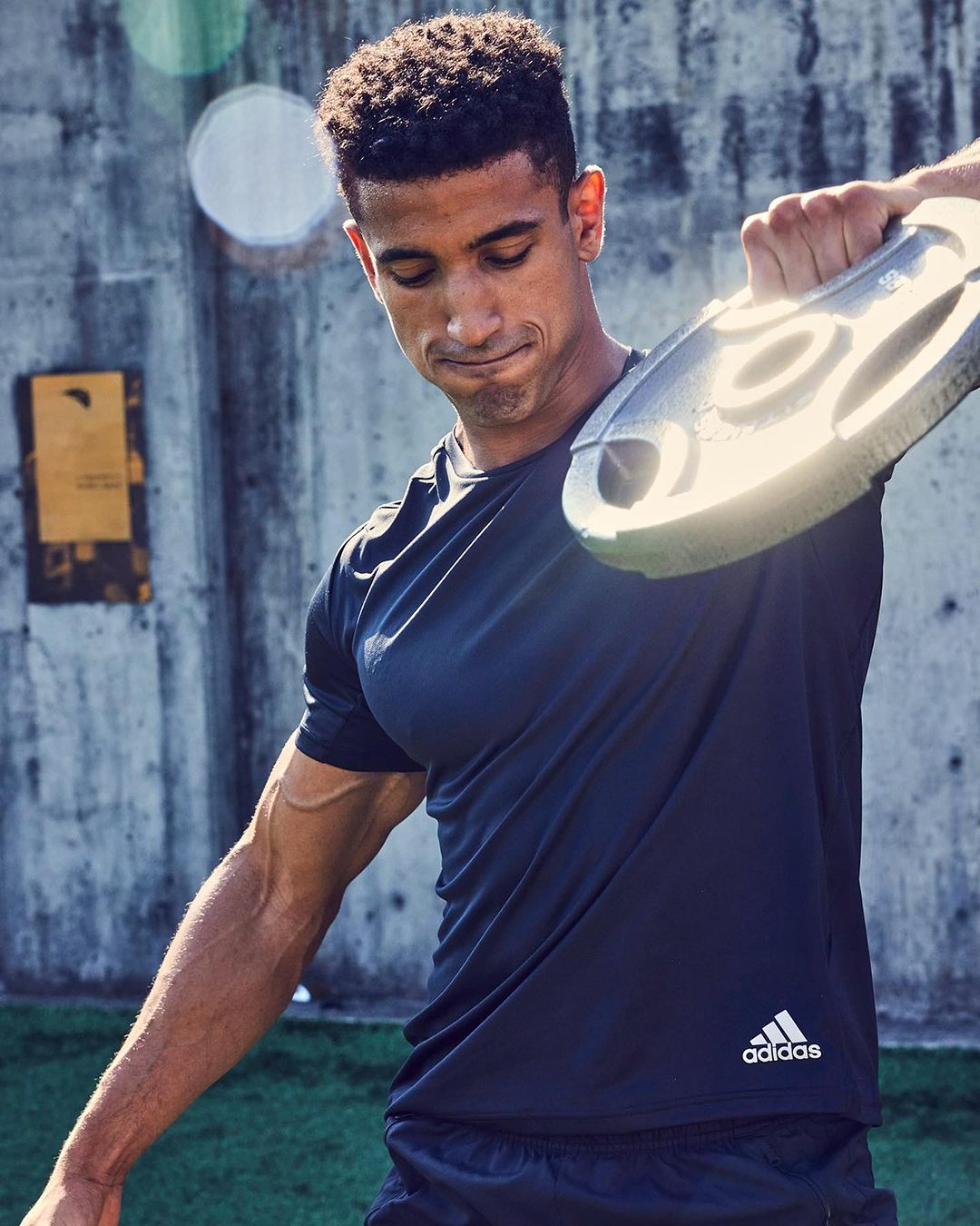 Athletic man works out outdoors with a gray weight plate, wearing dark athletic wear with an Adidas logo, against a concrete wall.