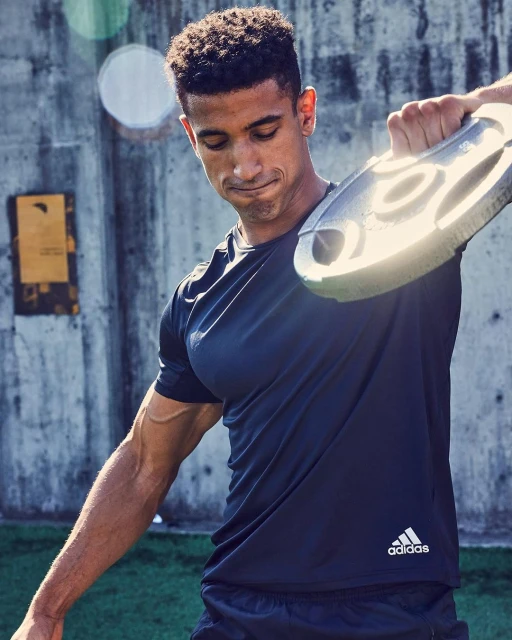 Athletic man works out outdoors with a gray weight plate, wearing dark athletic wear with an Adidas logo, against a concrete wall.
