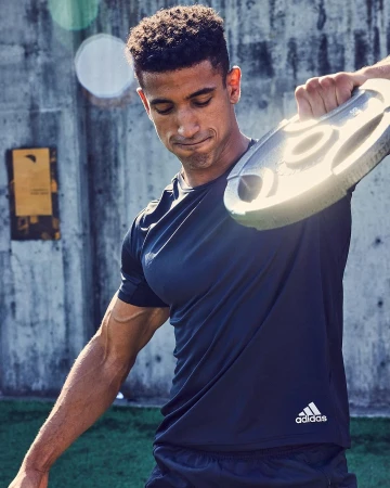 Athletic man works out outdoors with a gray weight plate, wearing dark athletic wear with an Adidas logo, against a concrete wall.
