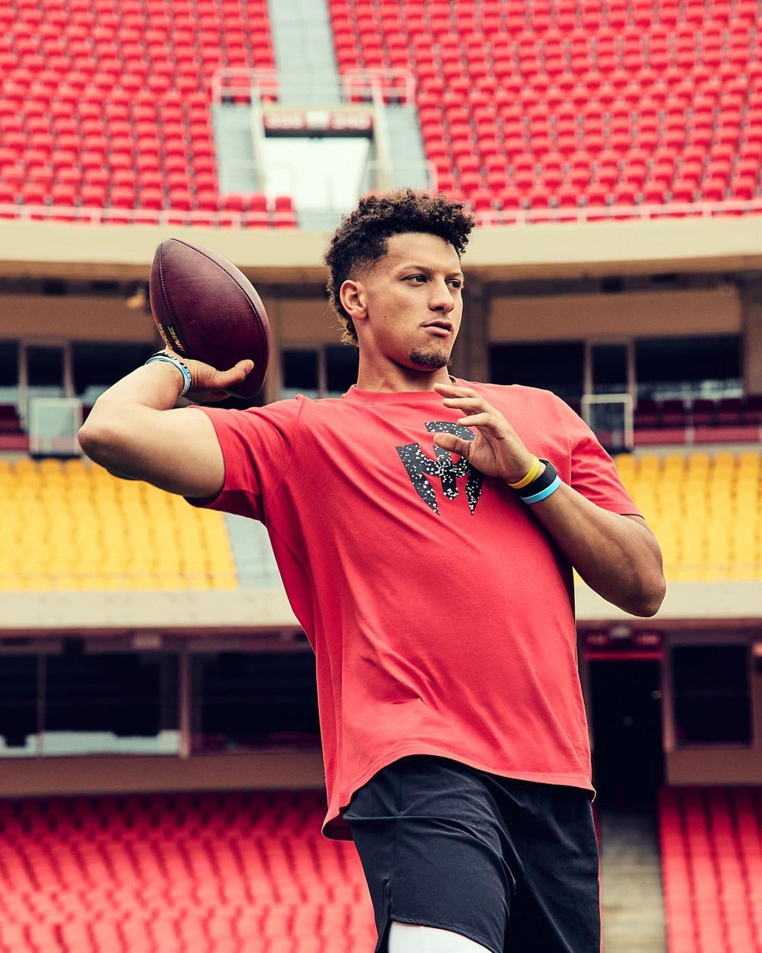 Patrick Mahomes throws a football in a stadium, wearing a red t-shirt and black shorts, while standing in front of empty red seats.