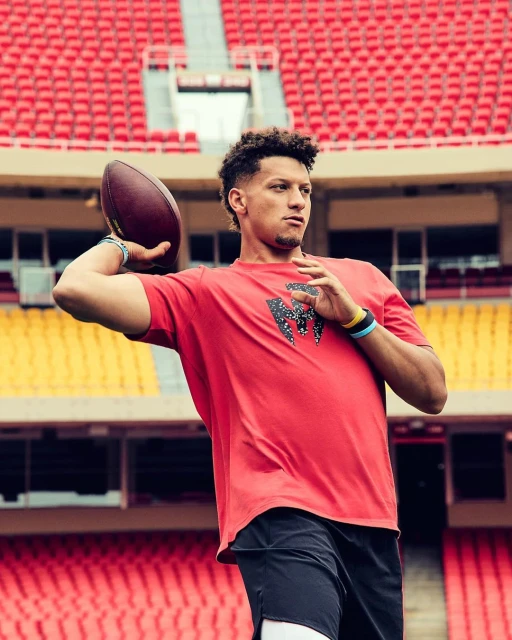 Patrick Mahomes throws a football in a stadium, wearing a red t-shirt and black shorts, while standing in front of empty red seats.