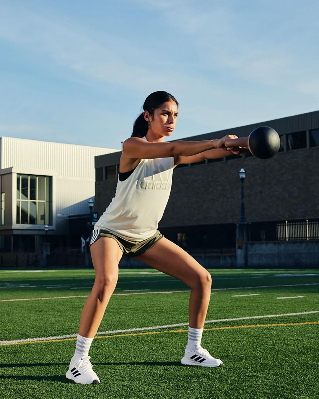 A woman in athletic wear performs a kettlebell exercise outdoors on a green field, with buildings and sky in the background.