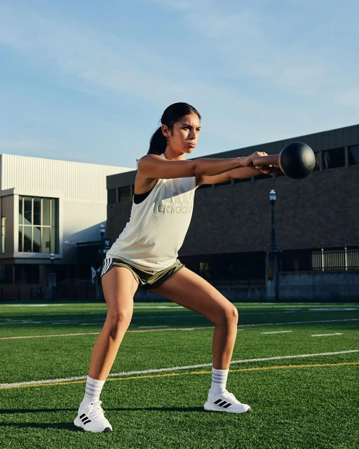 A woman in athletic wear performs a kettlebell exercise outdoors on a green field, with buildings and sky in the background.