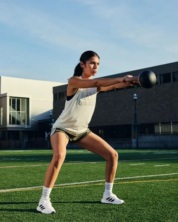 A woman in athletic wear performs a kettlebell exercise outdoors on a green field, with buildings and sky in the background.