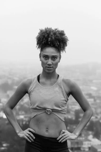A woman in workout gear stands outdoors against a hazy urban background, hands on hips and looking directly at the camera in black and white.