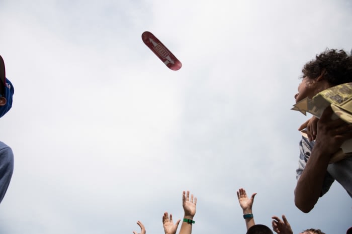 Skateboard suspended in air over group of people reaching up with their hands in outdoor daytime setting under partly cloudy sky.
