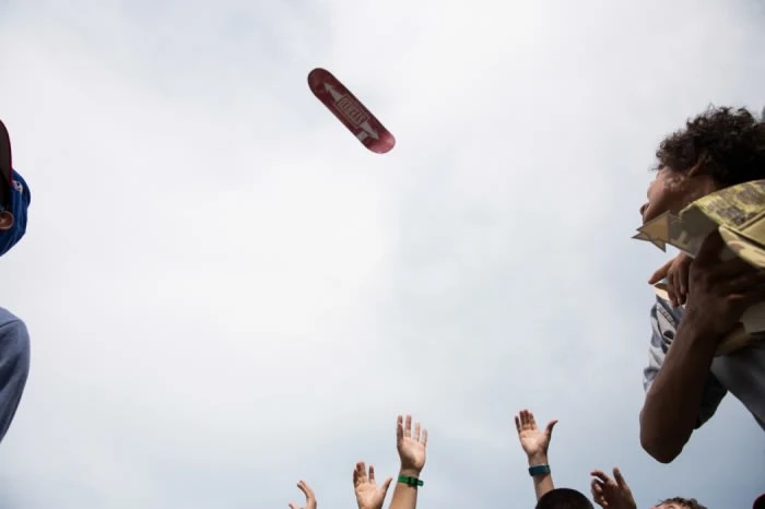 Skateboard suspended in air over group of people reaching up with their hands in outdoor daytime setting under partly cloudy sky.