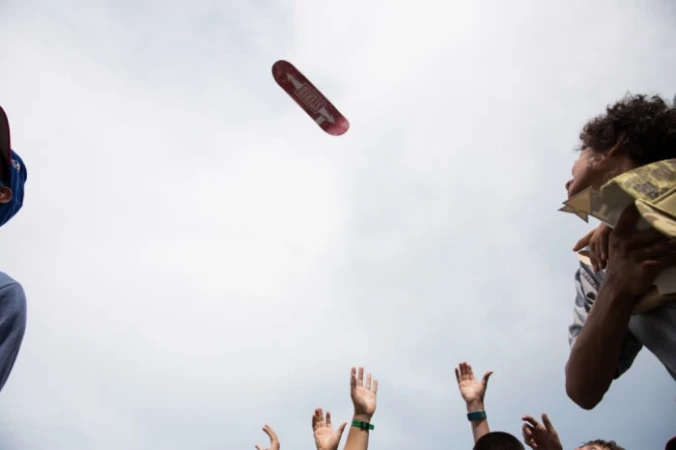 Skateboard suspended in air over group of people reaching up with their hands in outdoor daytime setting under partly cloudy sky.
