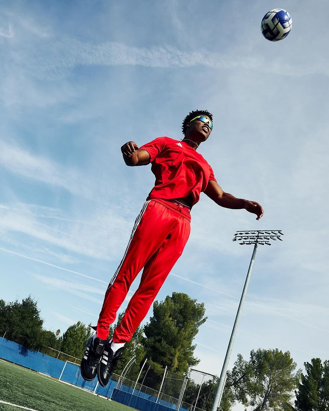 An adult male in red sportswear leaps on a grassy field, heading a soccer ball beneath a blue sky with light clouds and stadium lights.