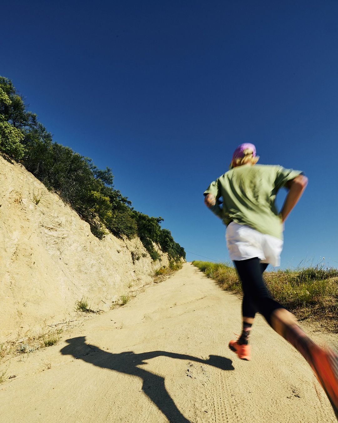 Woman trail running outdoors on a dirt path under a clear blue sky, captured in a sunny, wide shot, focused on fitness and nature.
